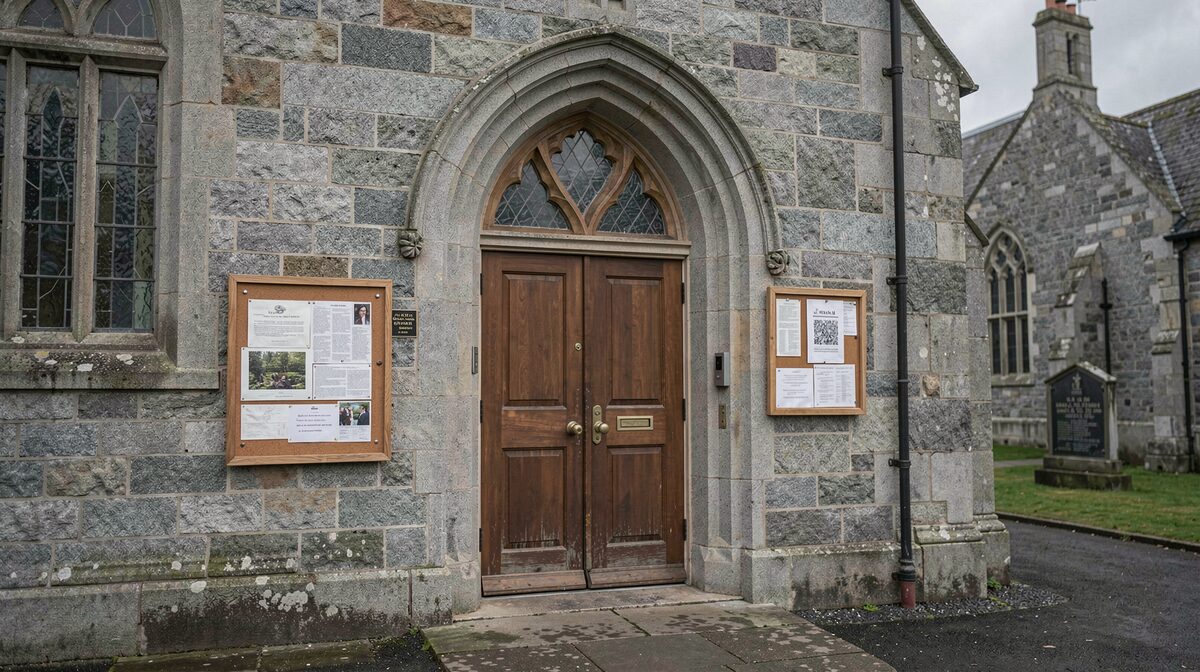 The west door of a grey granite Scottish Episcopal church on an overcast afternoon, a notice board beside the door filmed straight on with soft shadowless light.